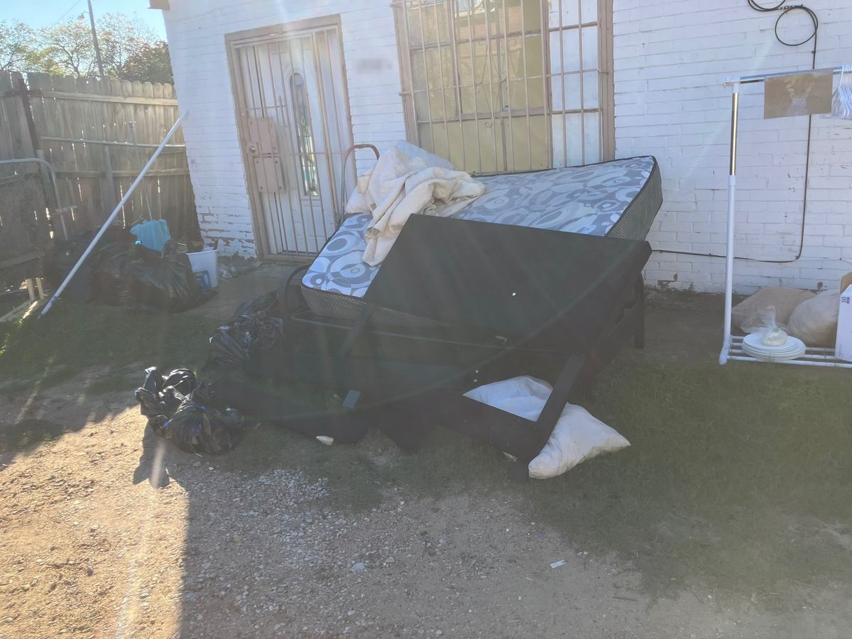 Mattress, bed frame, pillows and trash bags stacked outside a small brick house
